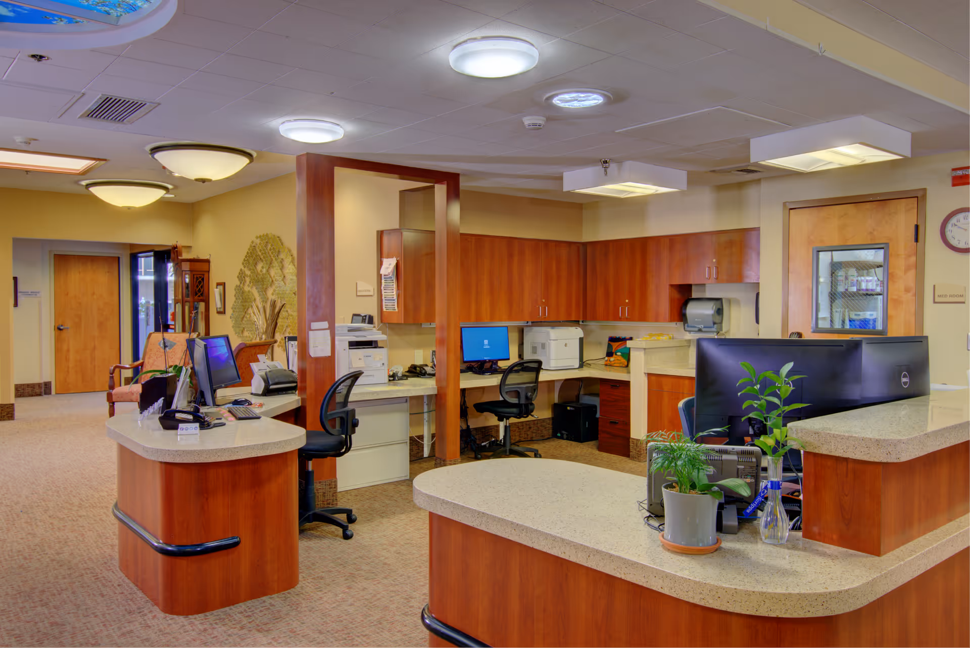 Reception area inside St. Paul's Skilled Nursing and Rehabilitation facility with a curved front desk, computer workstations, office chairs, wooden cabinetry, and potted plants. The space is well-lit with ceiling lights and has a carpeted floor. A hallway with wooden doors and seating is visible in the background.