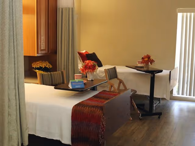 A room with two hospital-style beds separated by curtains. Each bed has a small overbed table with flowers and a welcome sign. The room has wooden flooring and vertical blinds covering a window or door.