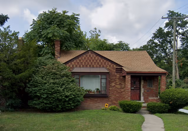 A single-story brick house with a brown shingled roof, a chimney, and a small front porch with a red door. The house is surrounded by green bushes and trees, with a concrete pathway leading to the front door. The sky is partly cloudy.