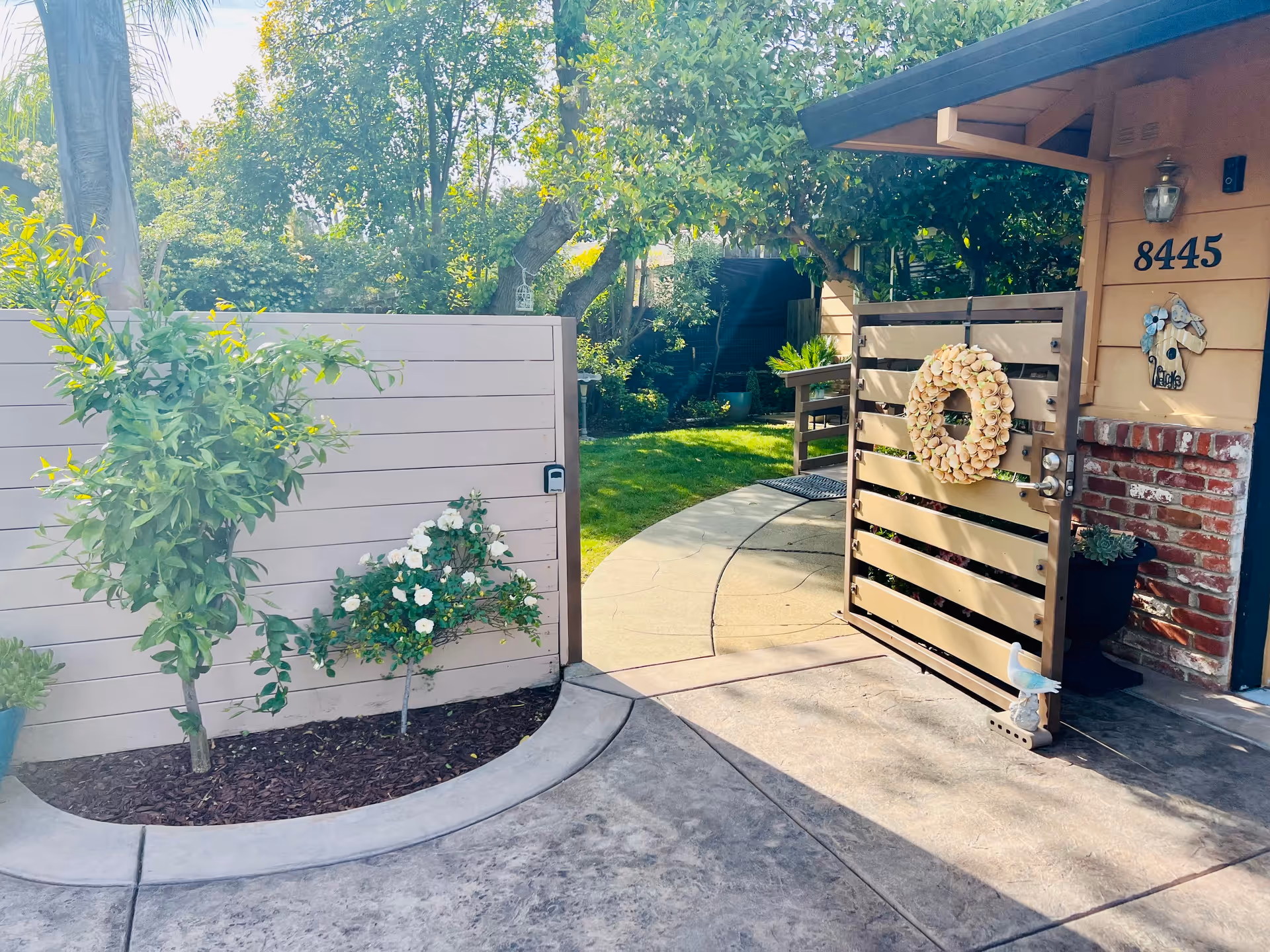 An open wooden gate with a decorative wreath leads to a garden area with green grass, trees, and plants. The gate is attached to a beige wall and a house with the number 8445 displayed. There is a small bird statue on the ground near the gate and a brick wall section on the house exterior.