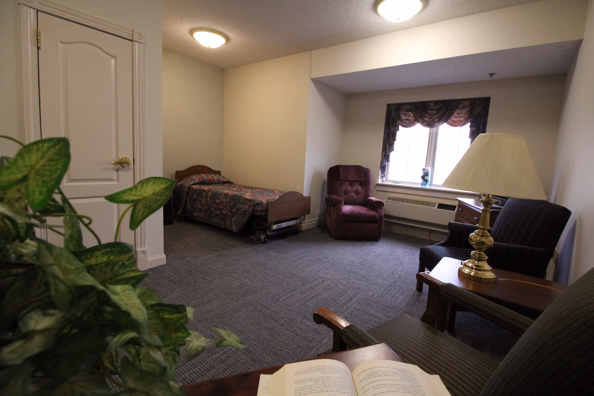 A small, simple bedroom in a senior living facility with a single bed covered in a patterned bedspread, a burgundy recliner chair near a window with floral curtains, a wooden side table with a brass lamp, and two upholstered chairs. A green leafy plant is partially visible in the foreground, and an open book lies on a table.