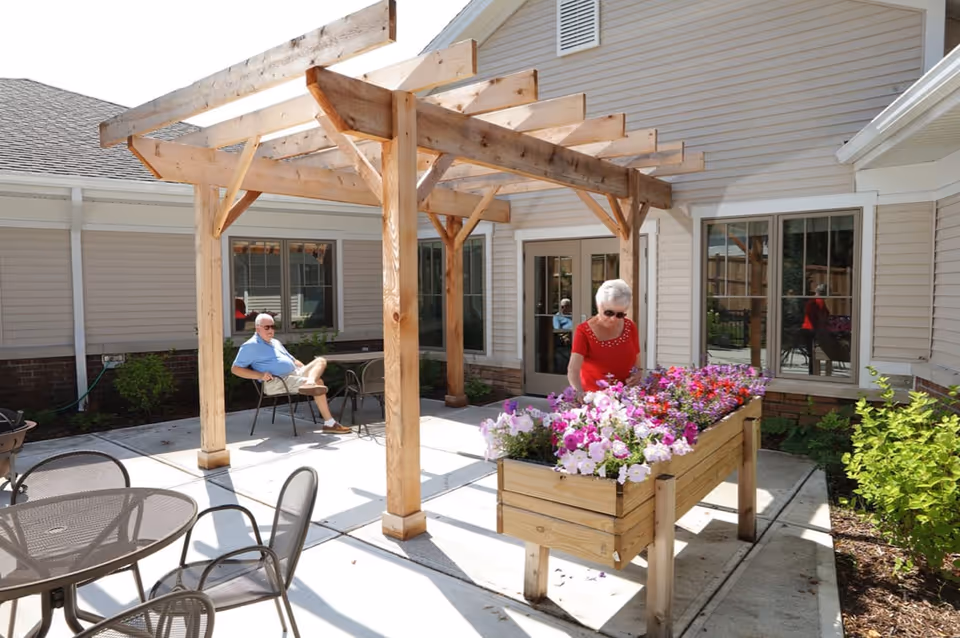 An outdoor patio area at Resthaven Maple Woods Cottages featuring a wooden pergola, metal patio tables and chairs, and a raised wooden planter box filled with colorful flowers. An elderly woman in a red shirt is tending to the flowers, while an elderly man in a blue shirt and shorts sits on a chair under the pergola. The building exterior is beige with white trim and multiple windows.