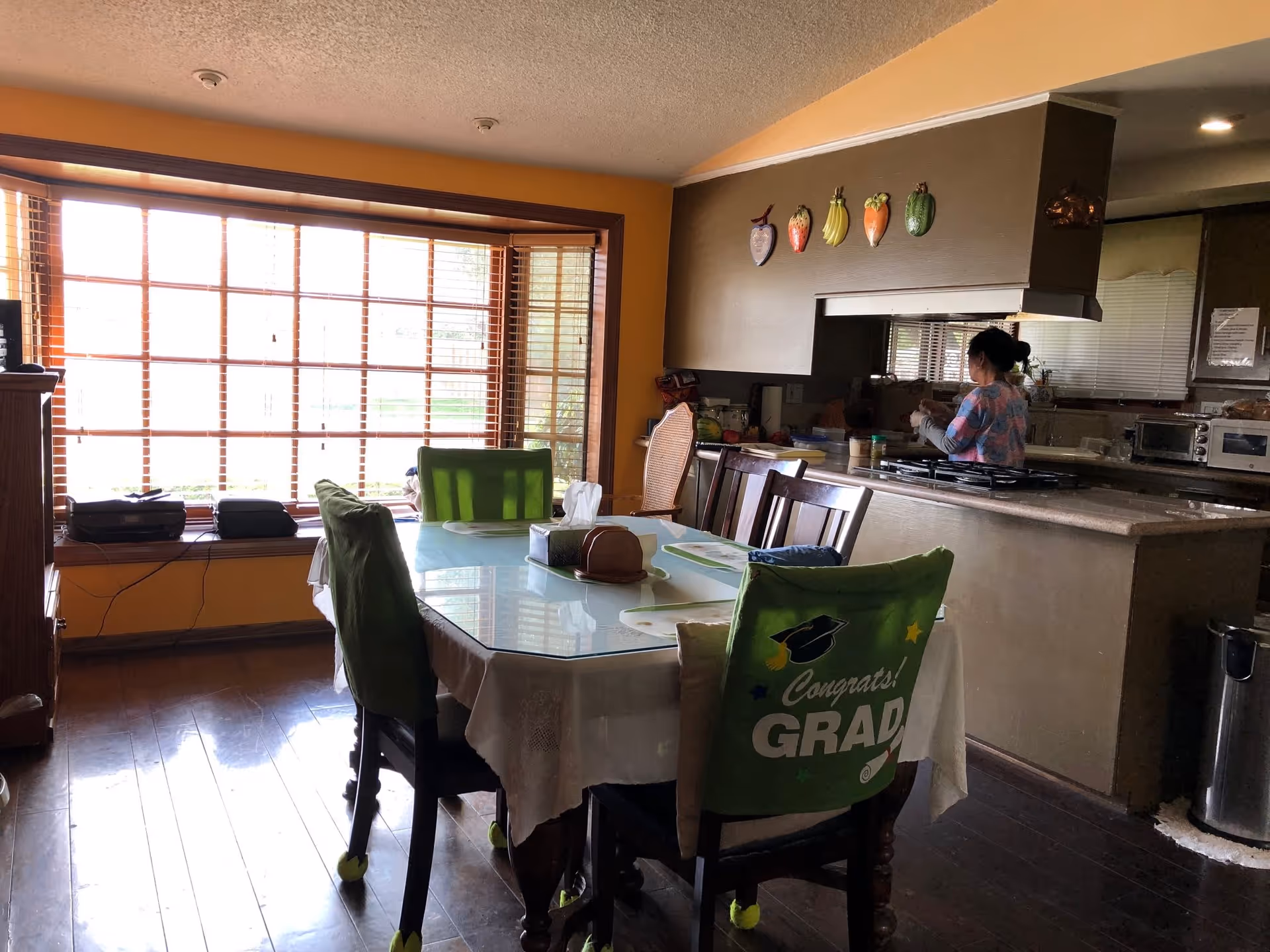 Dining area with a glass-topped table and green chair covers by a large bay window, open to a kitchen where a person stands at the counter.