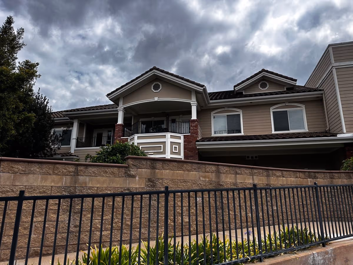 Front exterior of a two-story beige retirement community building with balconies, a stone retaining wall and iron fence under a cloudy sky.