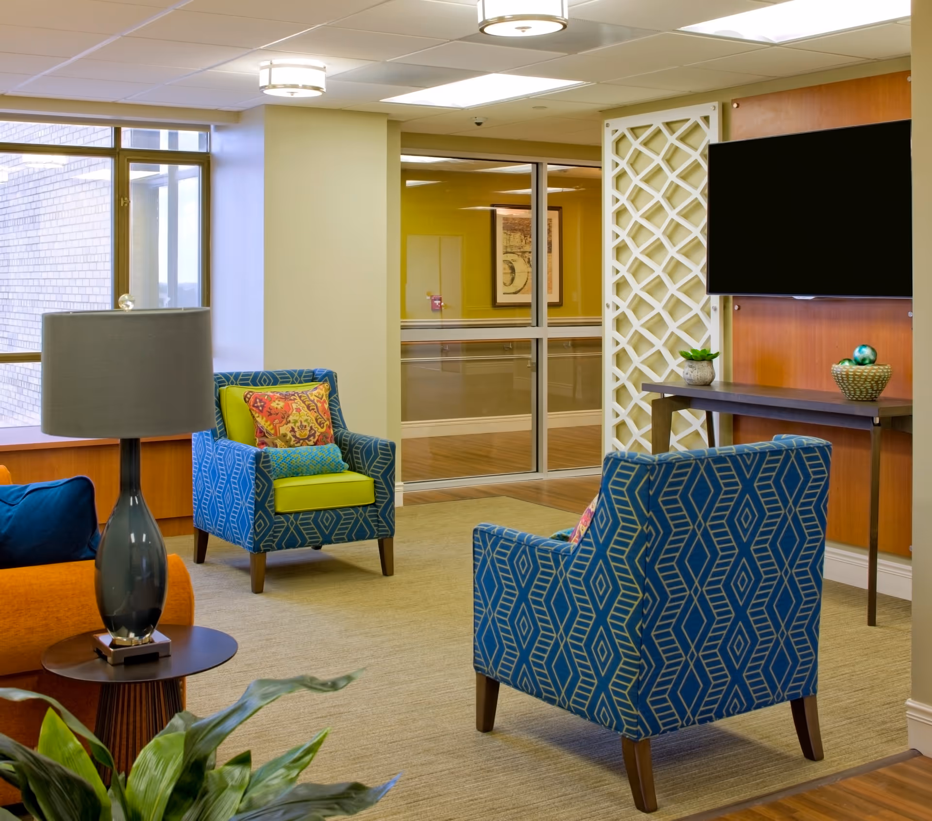 Bright lounge area with blue patterned armchairs, a wall TV, lattice divider, and glass doors.