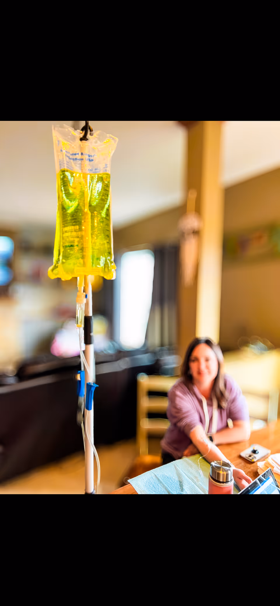 Intravenous infusion bag in foreground with a seated resident in a cozy living room.