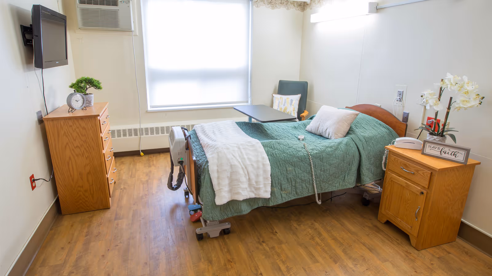 A single-occupancy senior living bedroom with a green bedspread, wooden dressers, a wall-mounted TV, and a window.