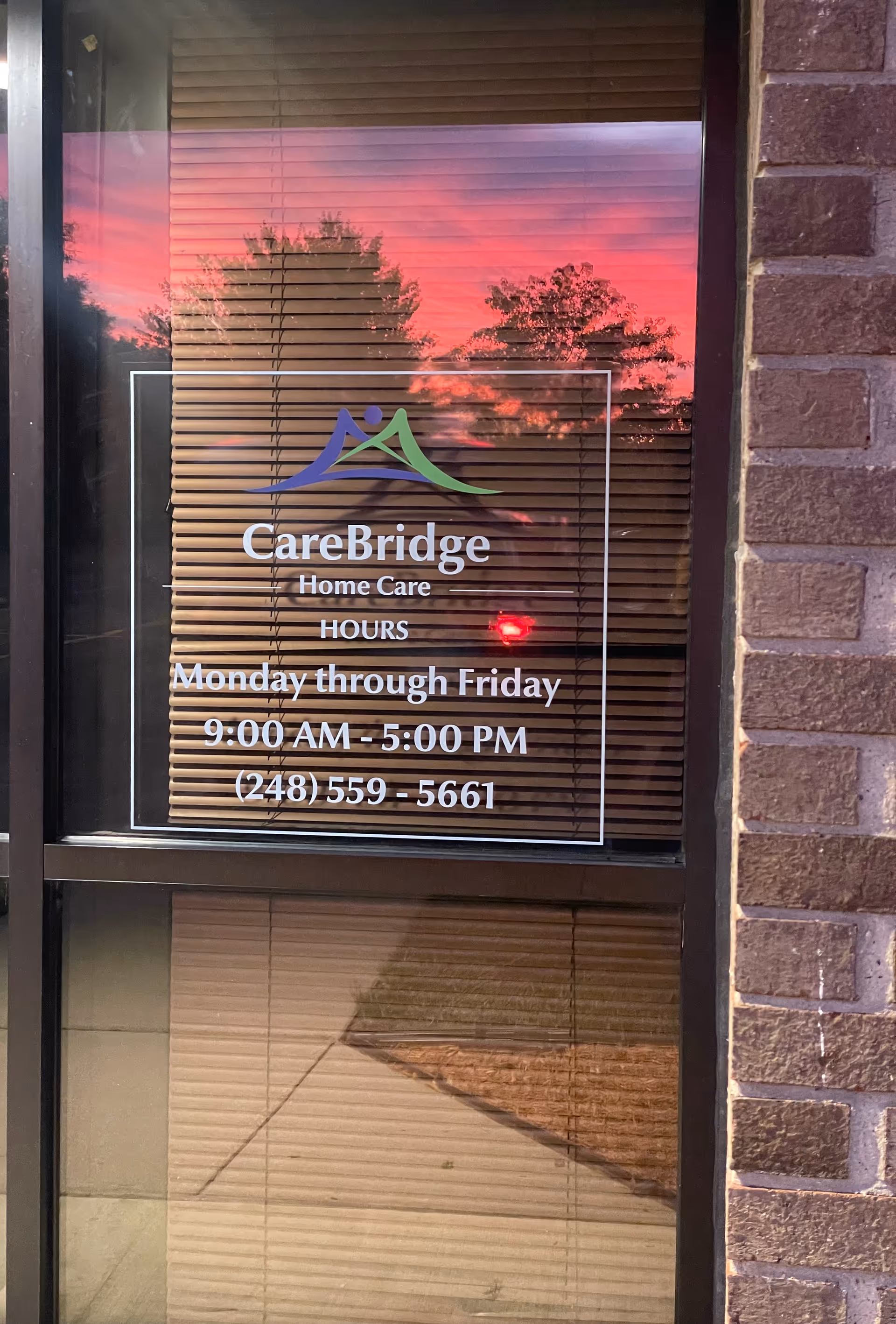 Glass storefront window displays CareBridge Home Care sign with sunset reflection on blinds.