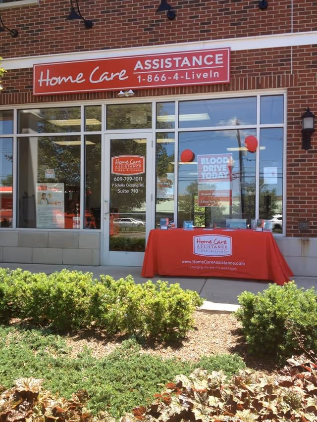 Brick storefront of Home Care Assistance Plainsboro Senior Care, glass doors, and a red promotional table.