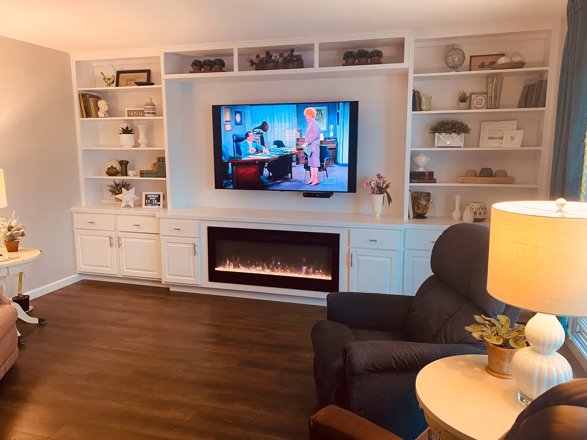 A cozy living room with built-in white shelves and cabinets surrounding a wall-mounted flat-screen TV showing a vintage scene. Below the TV is a modern electric fireplace with flames. The room has dark wood flooring, a comfortable dark recliner chair, a round side table with a white lamp and a small potted plant. The shelves are decorated with various plants, books, and decorative items.
