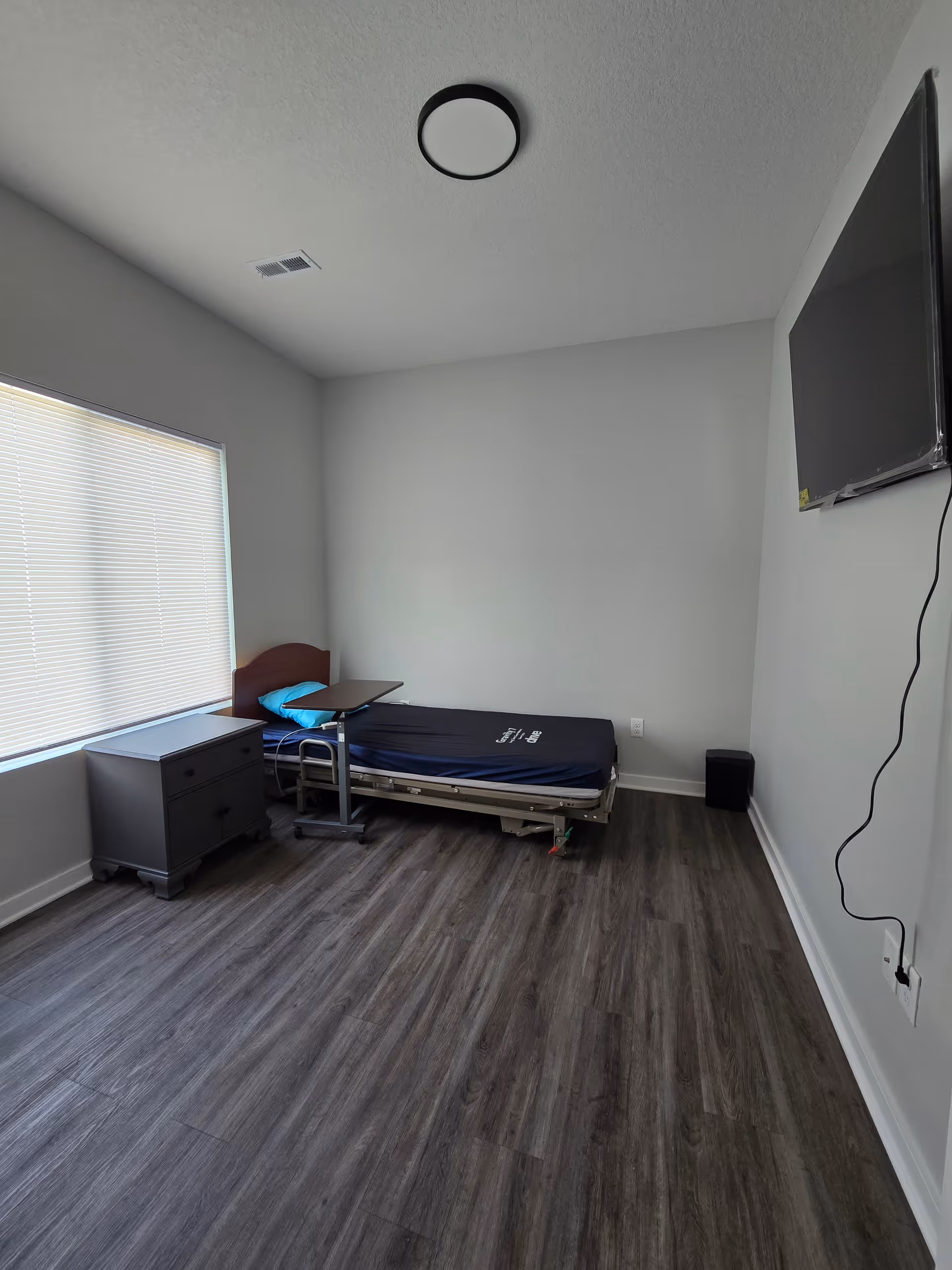 A simple bedroom with a single hospital-style bed with a blue pillow and a black mattress. Next to the bed is a small dark-colored nightstand and an adjustable overbed table. The room has wood-look flooring, a window with closed blinds, a ceiling light fixture, and a wall-mounted flat-screen TV with visible cords.