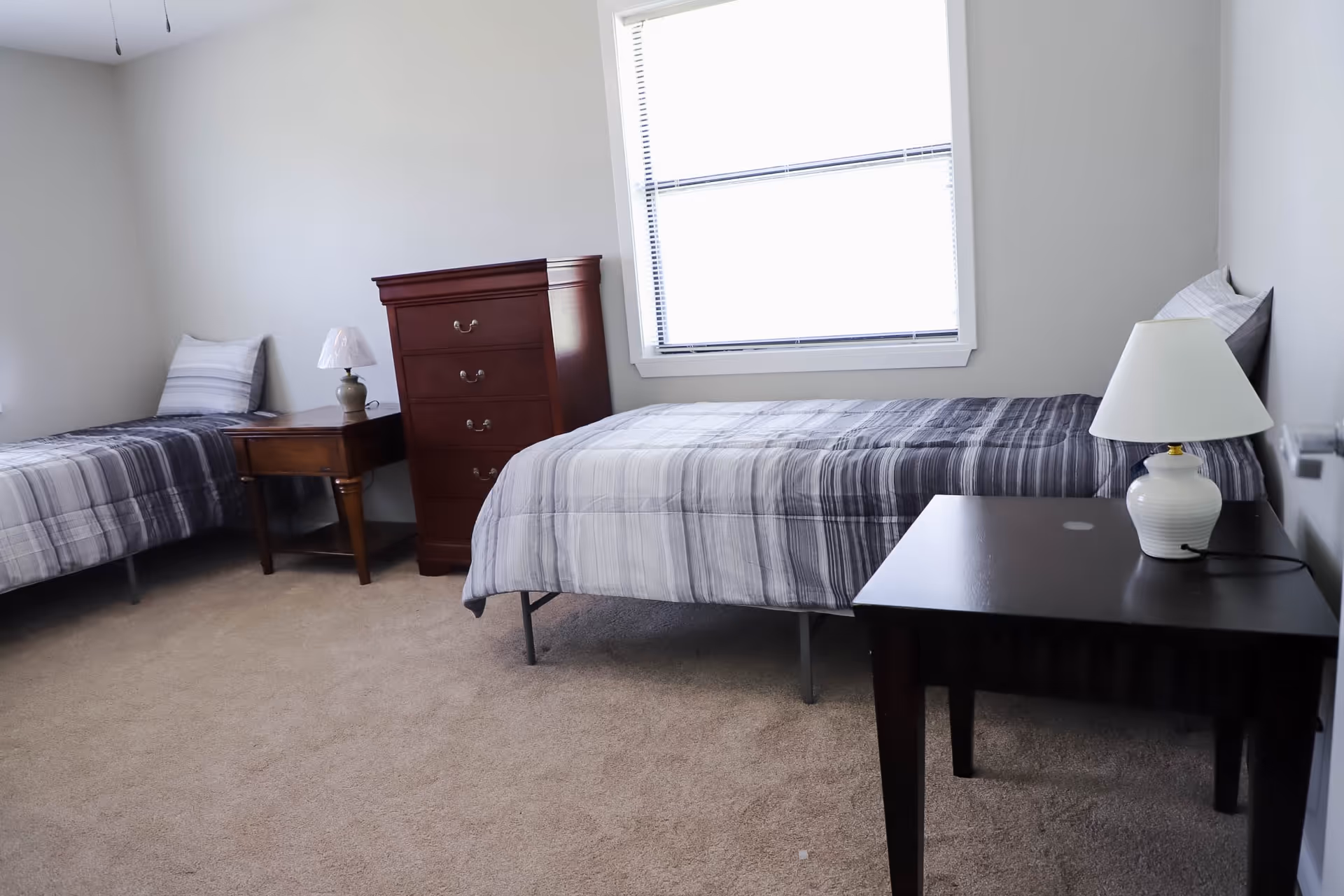A simple bedroom with two single beds, each with striped gray bedding and a pillow. Between the beds is a wooden chest of drawers and a small wooden nightstand with a lamp. Another black table with a white lamp is positioned near the bed on the right. The room has beige carpet and a window with blinds letting in natural light.