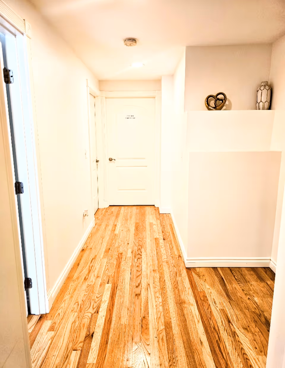 Bright interior hallway with hardwood floors, white walls, doors, and decorative shelf.