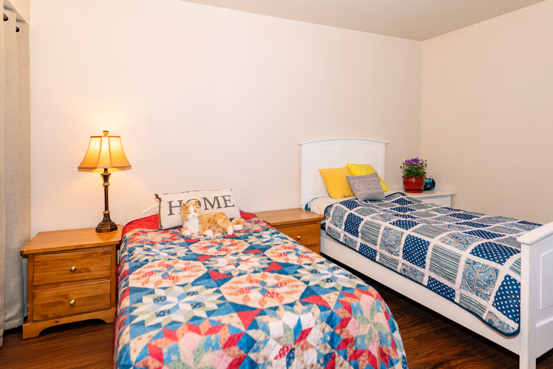 A cozy bedroom with two single beds, each with colorful patchwork quilts. The left bed has a pillow with the word 'HOME' and a stuffed animal on it. Between the beds is a wooden nightstand with a lamp. The right bed has yellow and gray pillows and a small white nightstand with a red pot containing purple flowers and a small blue clock.