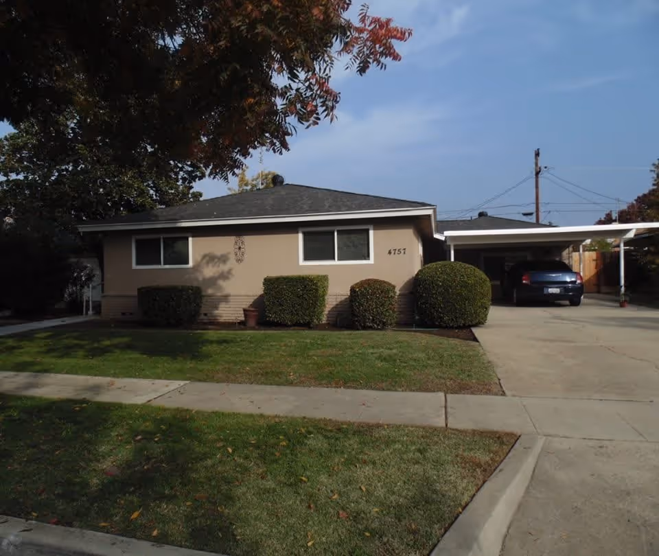 Single-story beige house with a driveway and carport, a parked car, front lawn and trimmed shrubs.