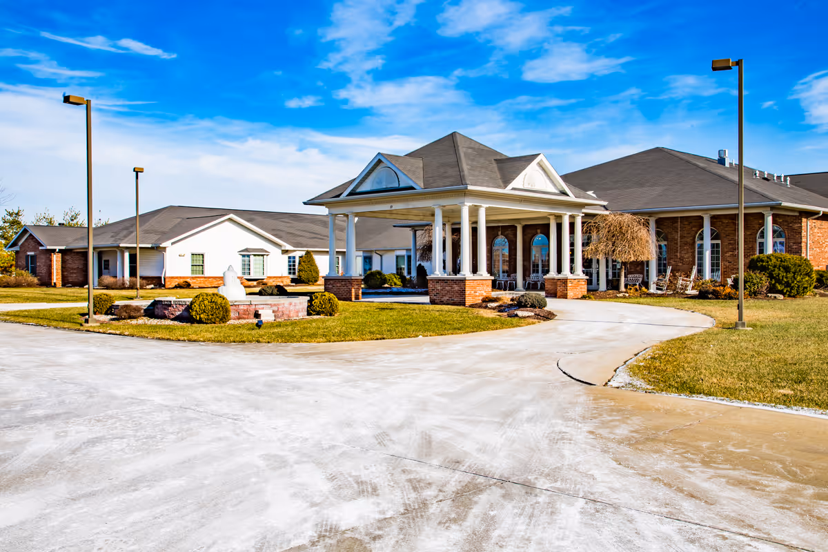 Exterior view of Charter Senior Living of Troy, showing a large building with a covered entrance supported by white columns, surrounded by a well-maintained lawn and a clear blue sky.