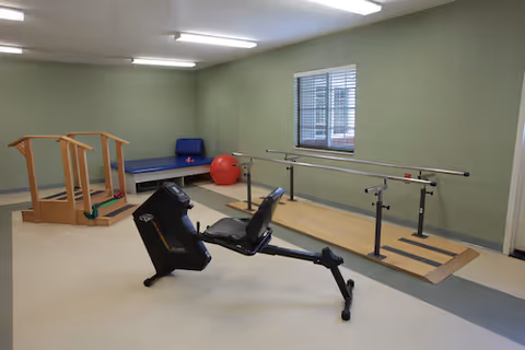 Small rehabilitation gym with parallel bars, exercise bike, ball, and padded mats inside a green-walled room.