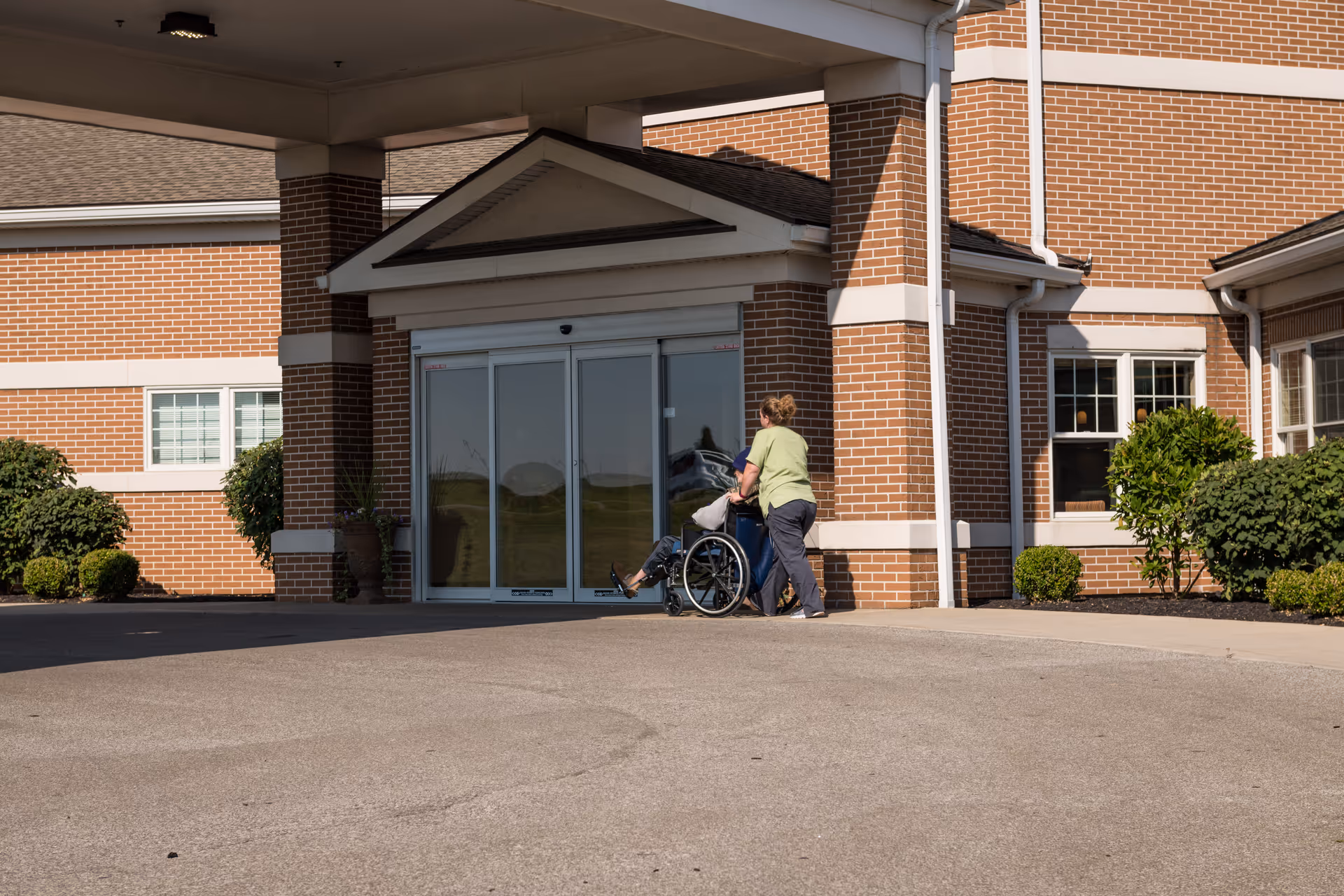 Two people near a wheelchair at the glass entrance of a brick senior living facility.
