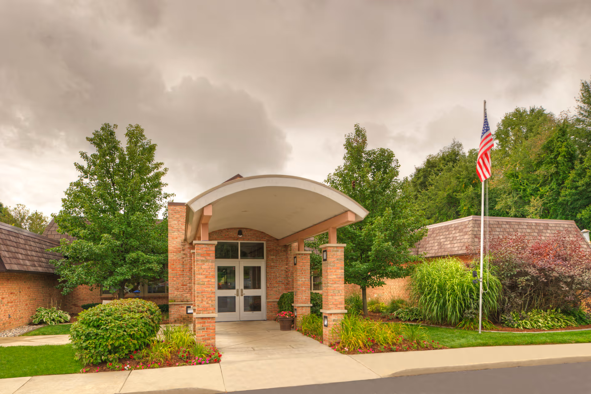 Entrance of a brick building with a covered walkway supported by columns, surrounded by green trees, bushes, and landscaped plants. An American flag is flying on a flagpole to the right of the entrance under a cloudy sky.