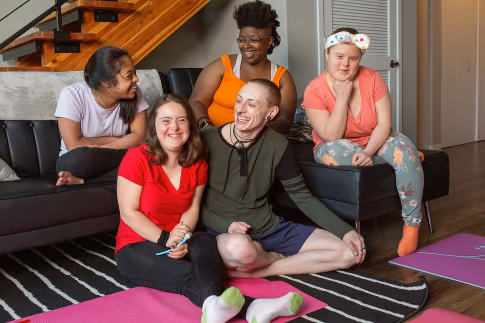 Group of smiling residents lounges together on a couch and mats in a bright living room.
