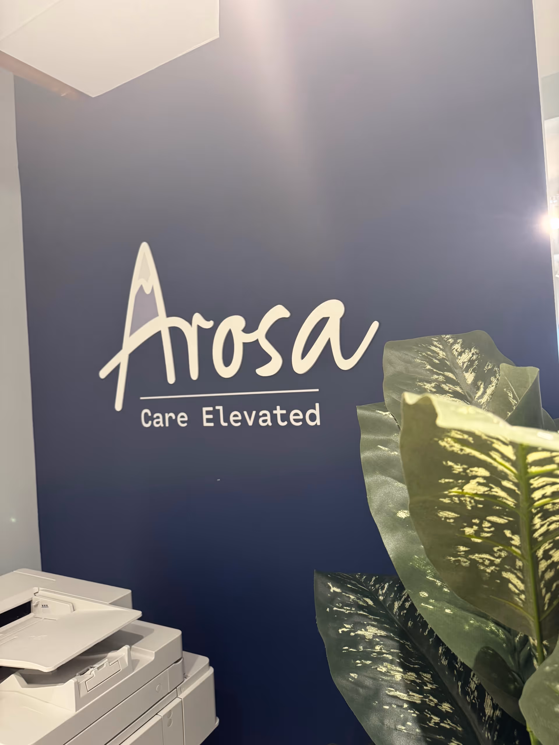 Blue wall displays Arosa logo with 'Care Elevated', printer on left, and a leafy plant.