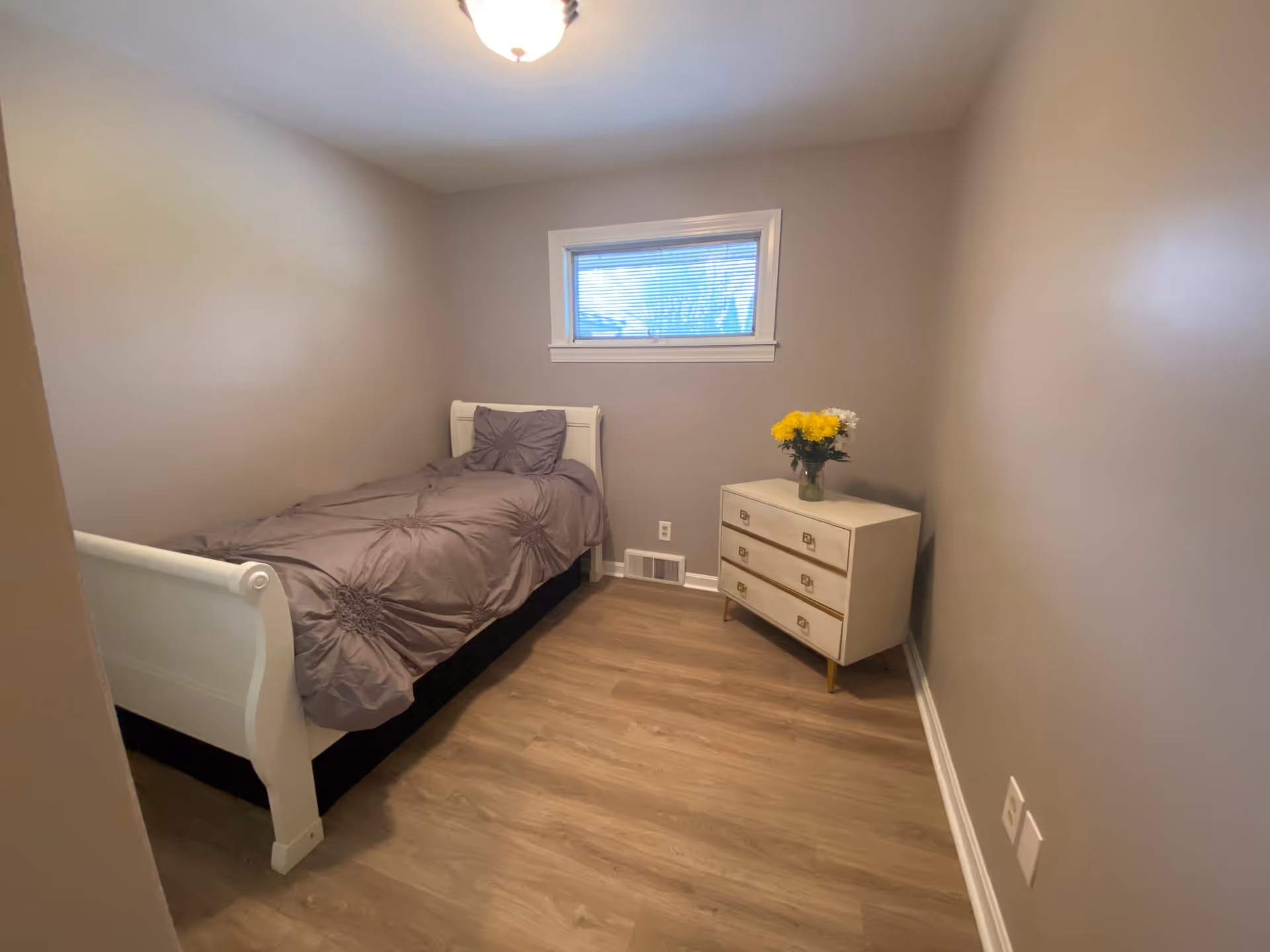 Small neutral-toned bedroom with a single bed, dresser, and a vase of yellow flowers.