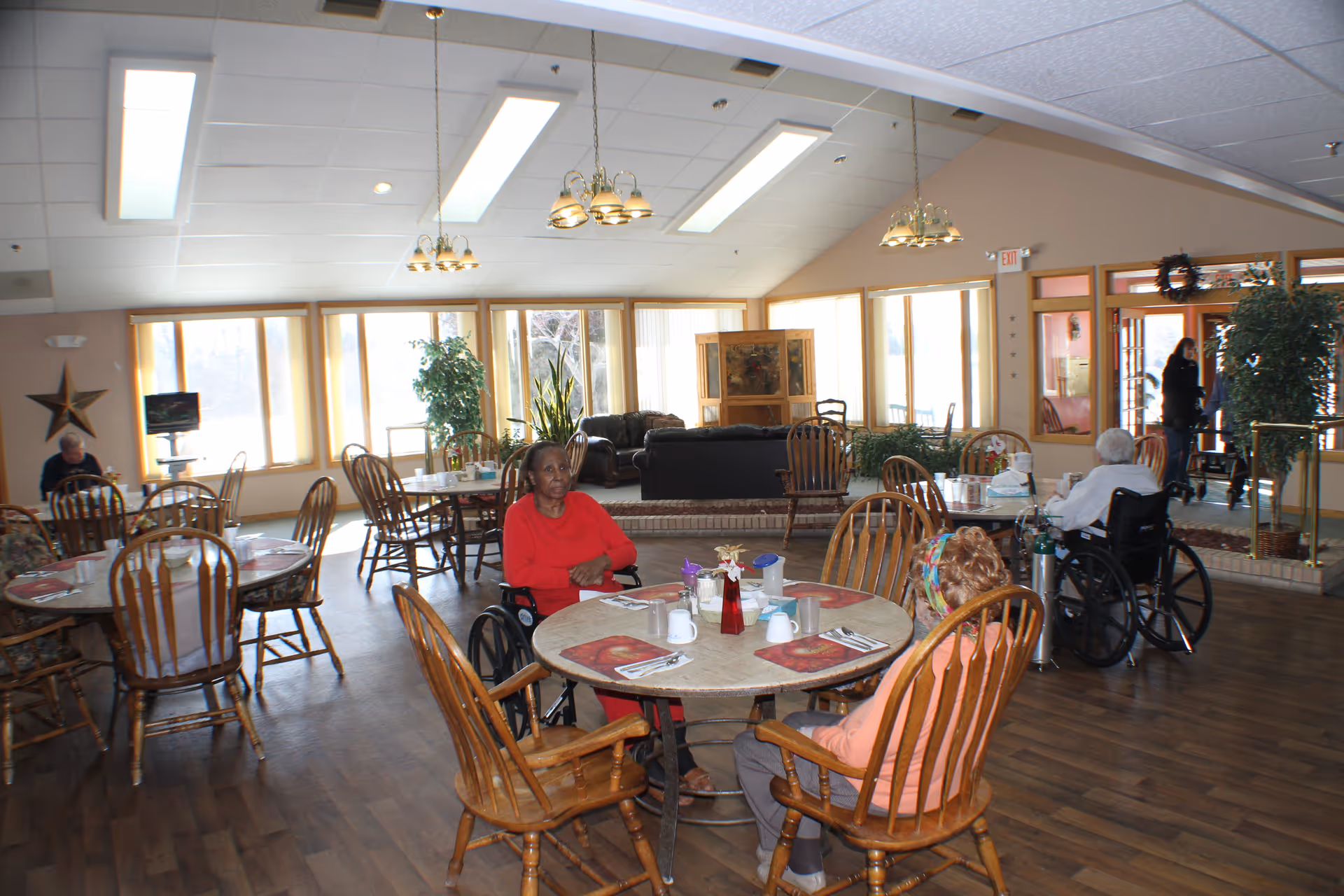 A bright dining room in a senior living facility with several round wooden tables and chairs. Three elderly individuals, two in wheelchairs, are seated at the tables. The room has large windows letting in natural light, hanging light fixtures, and a seating area with couches in the background. There are plants and decorative items around the room.