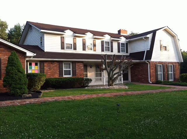 Two-story brick-and-white senior living house with a front porch and manicured lawn.