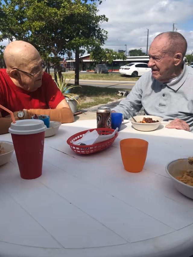 Two elderly men sitting at a white outdoor table having a meal together. One man is wearing a red shirt and glasses, and the other is wearing a light gray shirt. There are bowls of food, a red cup with a straw, a can of soda, and an orange cup on the table. Trees, a road, and parked cars are visible in the background under a partly cloudy sky.