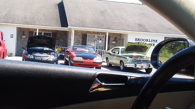 View through a car window of the Brookline Village and Cottages Senior Living entrance showing three cars with their hoods open parked in front of the building.