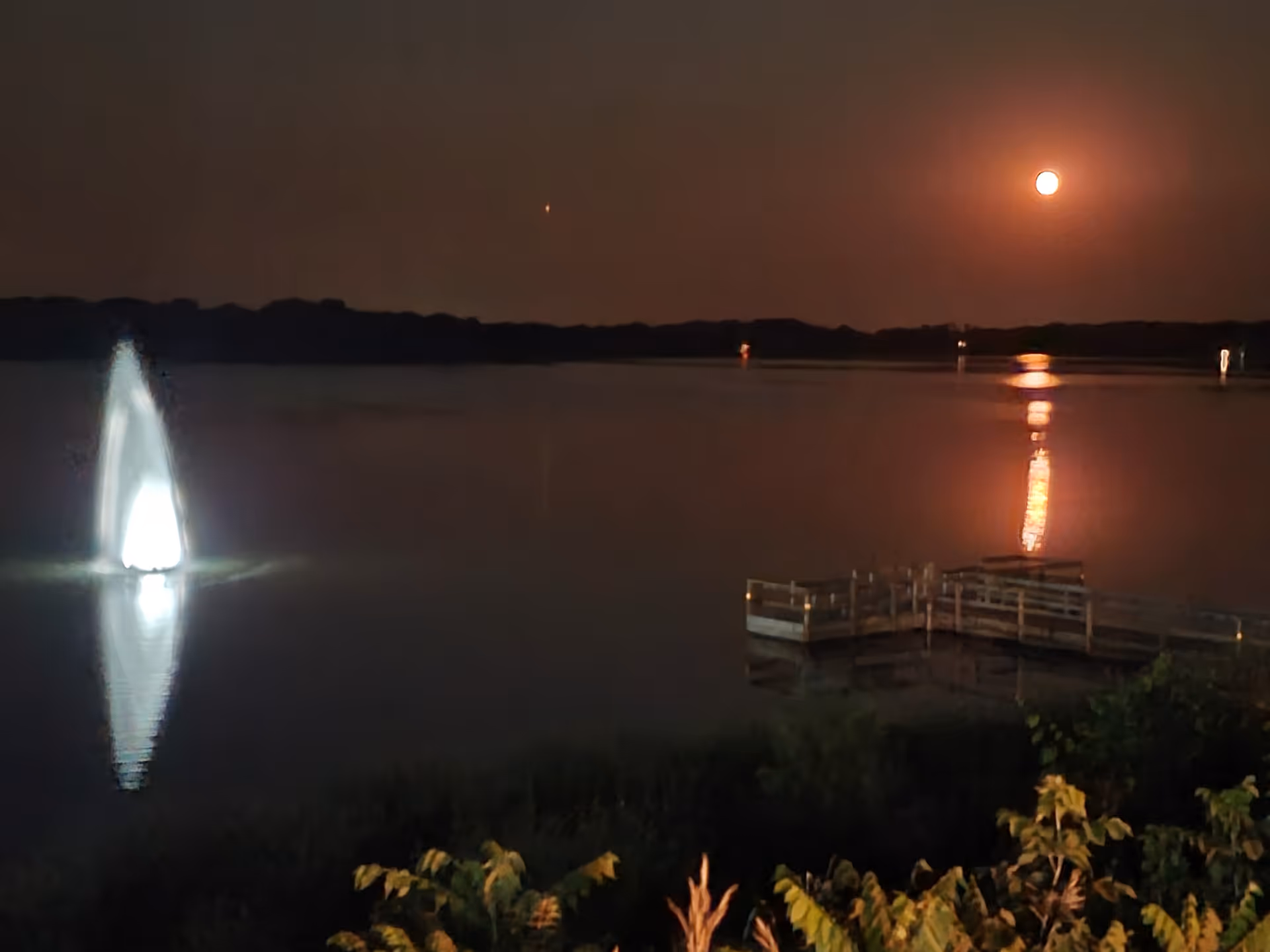 Night lakeside scene with illuminated fountain and wooden dock reflecting on water