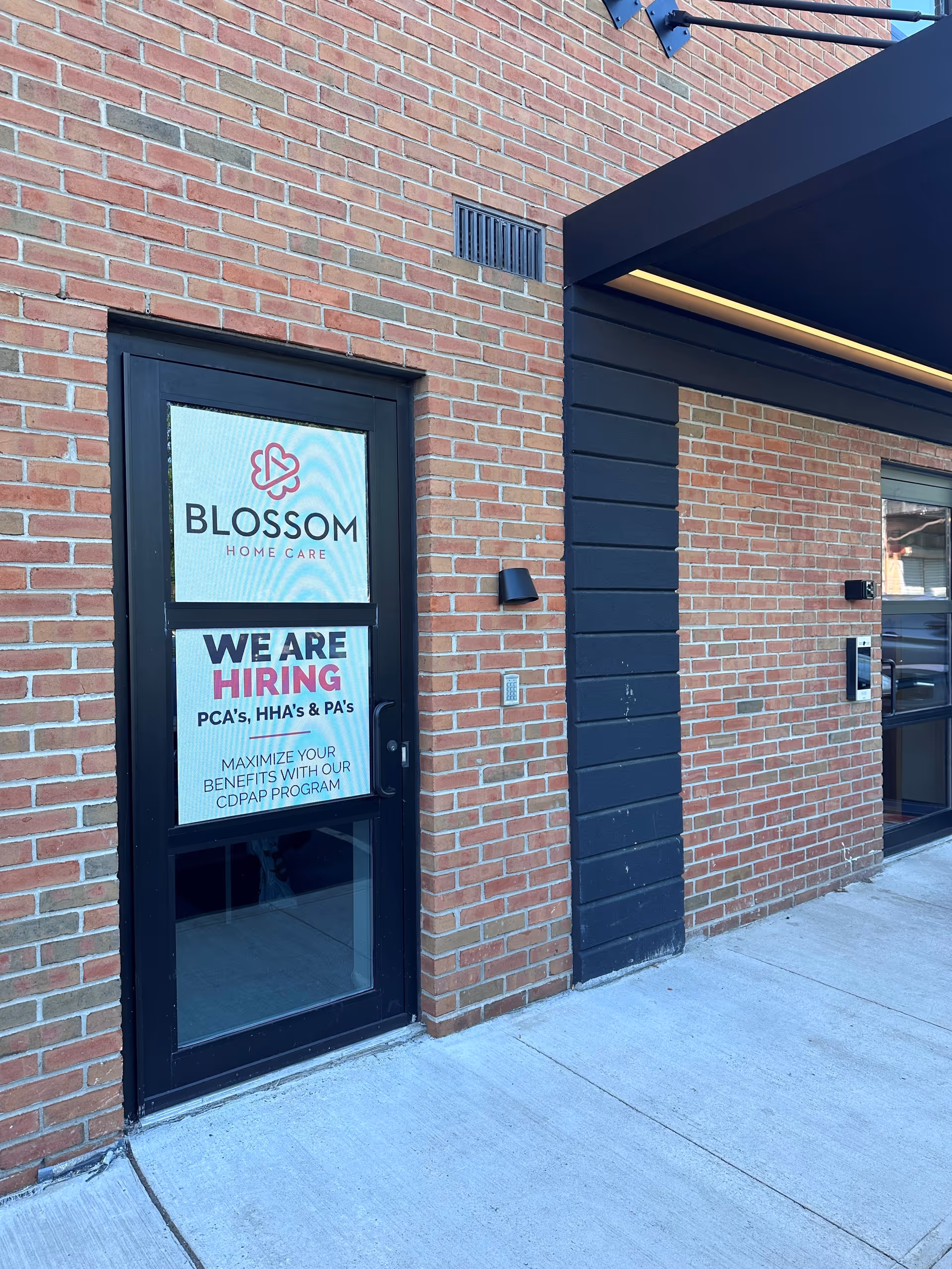 Brick exterior of Blossom Home Care with a glass door and We Are Hiring sign.