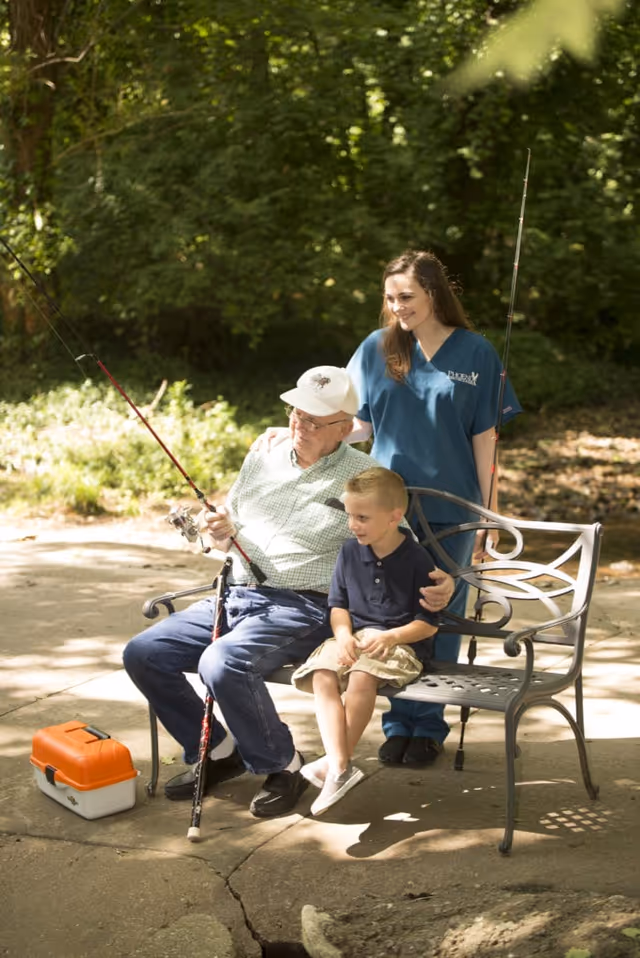 Elderly man fishes from a bench with a caregiver and child outdoors.