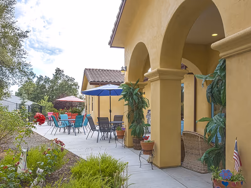 Outdoor patio area with yellow stucco arches, potted plants, and several tables with colorful umbrellas and chairs. There are green bushes and flowers along the walkway, and trees in the background under a partly cloudy sky.