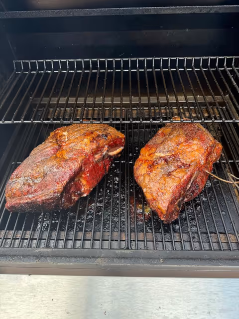 Two thick, smoked meat cuts cook on a metal barbecue grill.