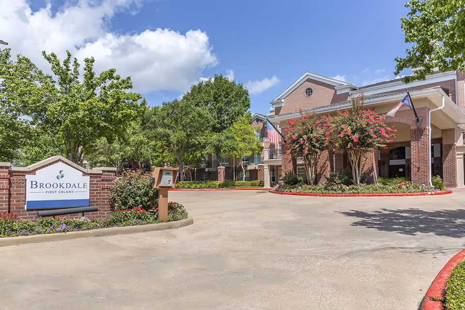Exterior view of Brookdale First Colony senior living facility showing the entrance driveway, a brick sign with the facility name, landscaped greenery, and a building with flags and flowering trees under a partly cloudy blue sky.