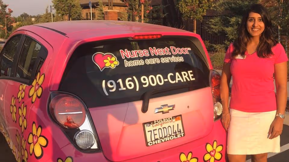 Smiling woman in pink top beside a pink Nurse Next Door van outdoors.
