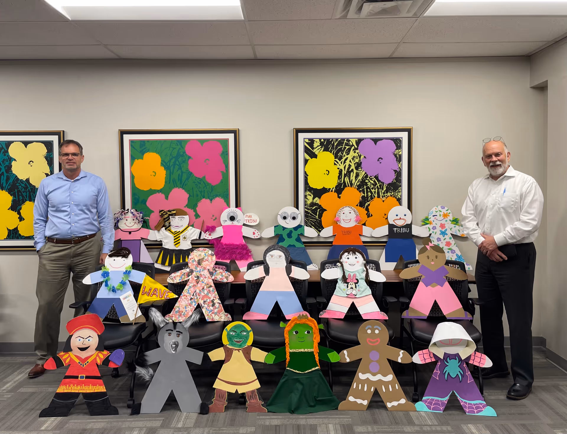 Two men stand beside numerous colorful handmade cutout figures arranged on chairs indoors.