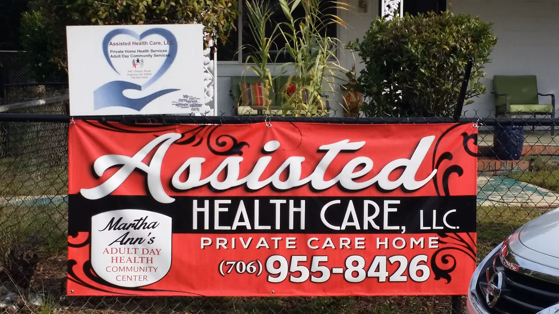 Red banner advertising Assisted Health Care LLC on a fence in front of a facility.