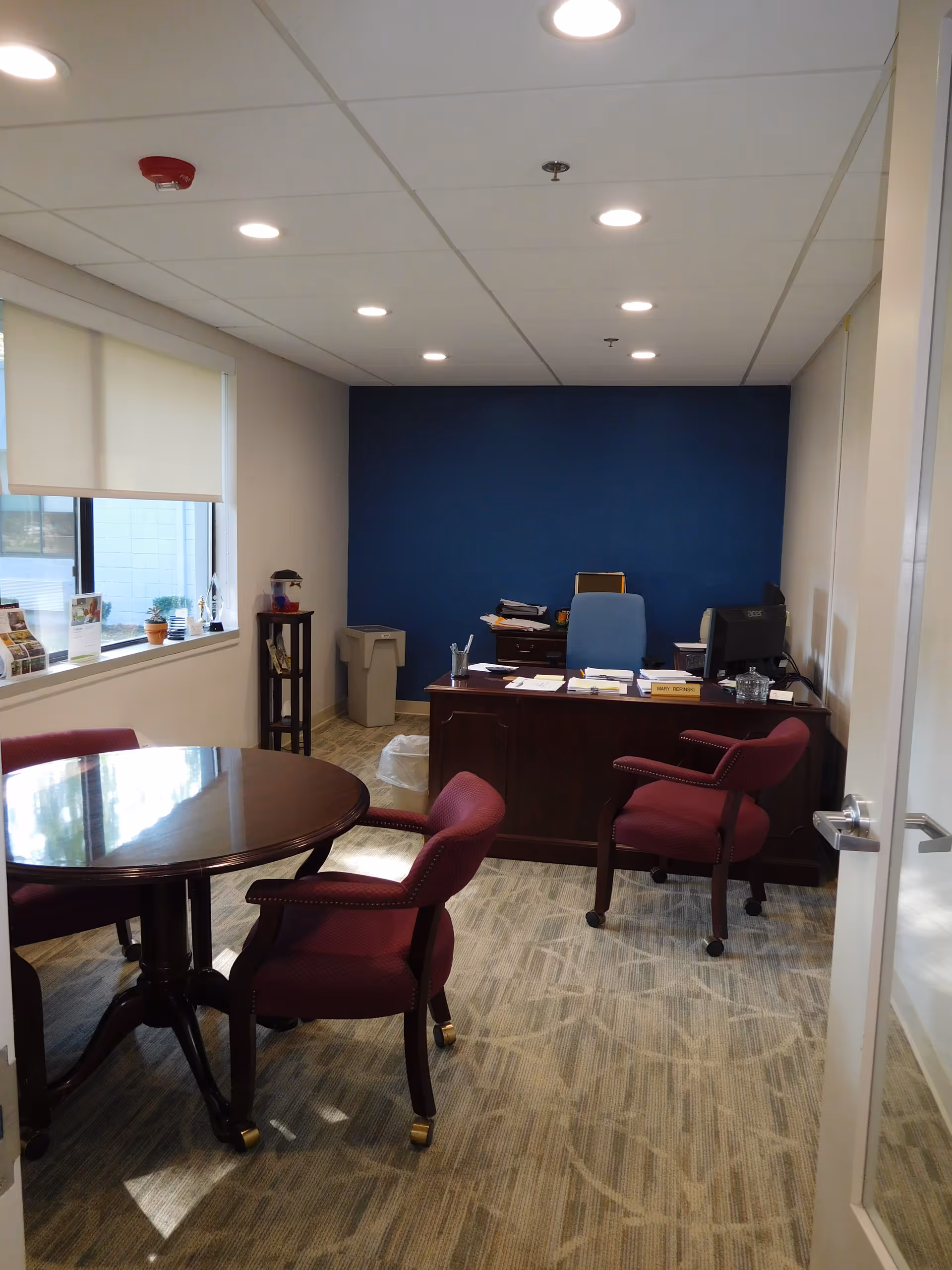Office interior with a round wooden meeting table and maroon chairs in the foreground and a desk against a blue accent wall at the back.