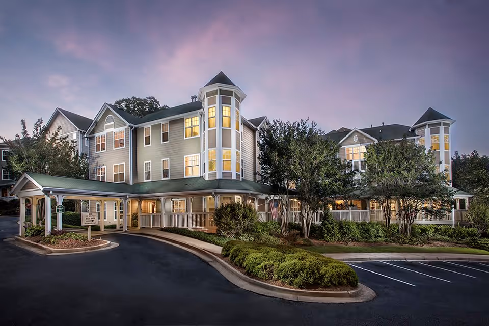 Exterior view of Winnwood Retirement Community building at dusk with lights on inside. The building has multiple stories, a covered entrance, and is surrounded by trees and landscaped bushes. The sky is purple and pink, indicating sunset.
