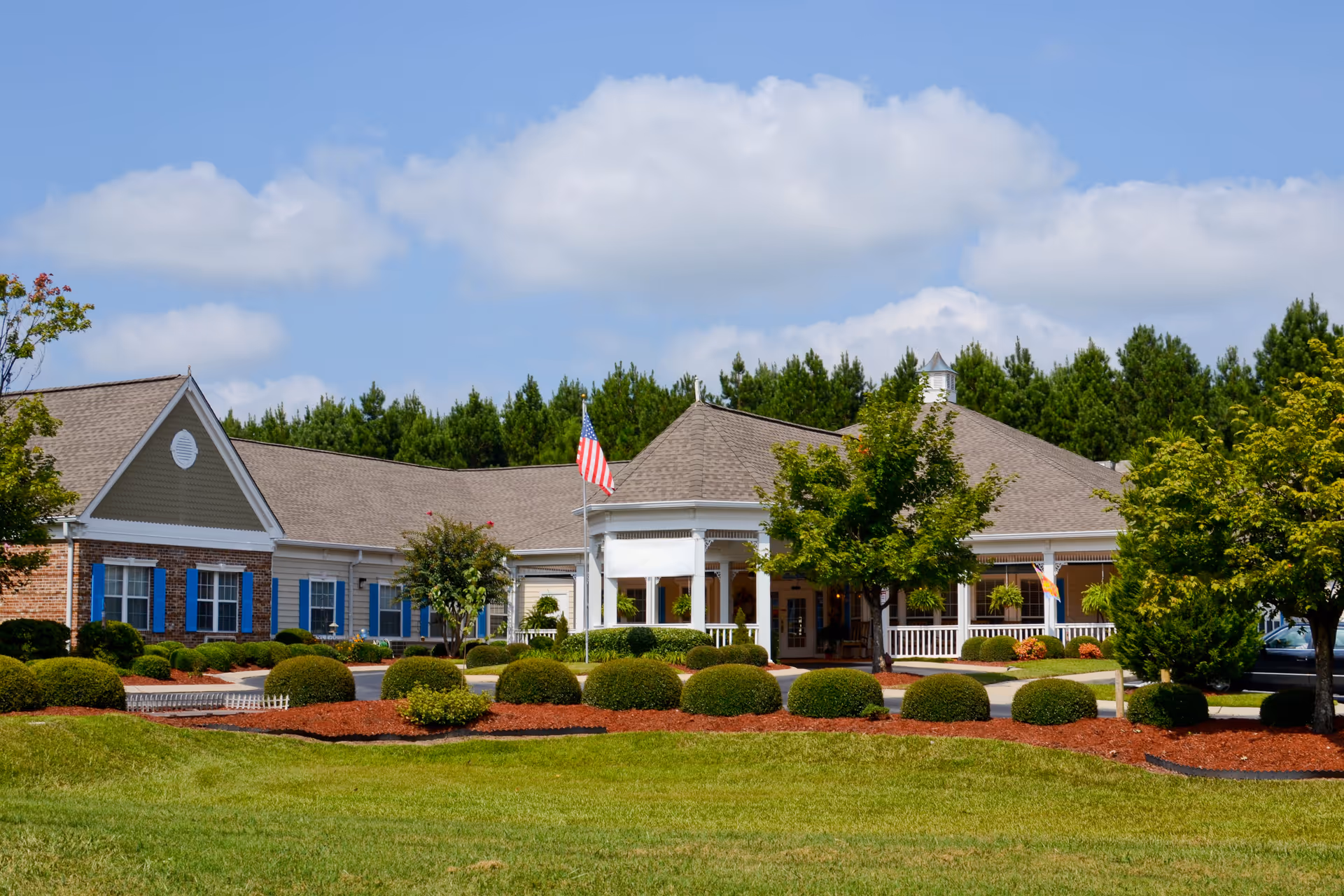 Exterior view of The Legacy of Orangeburg senior living facility showing a single-story building with a gabled roof, white columns, and a porch area. The building is surrounded by neatly trimmed bushes, trees, and a well-maintained lawn under a partly cloudy blue sky. An American flag is displayed near the entrance.