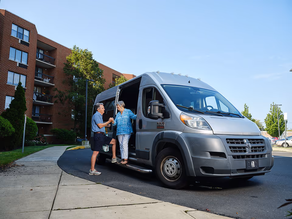 Two seniors exit a silver van outside a brick senior living building.