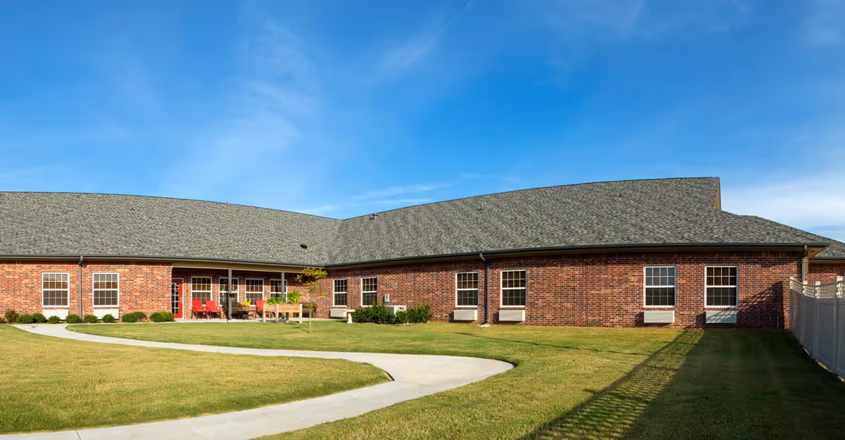 Exterior view of a single-story brick building with multiple windows and a gray shingled roof under a clear blue sky. There is a curved concrete walkway leading through a well-maintained grassy lawn with a few small bushes and outdoor seating with red chairs near the building.