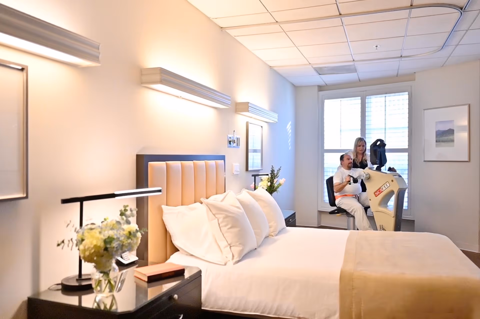 A well-lit bedroom in a healthcare facility with a neatly made bed featuring white pillows and a beige blanket. A bedside table holds a lamp and a vase with flowers. In the background, a man is seated on an exercise machine while a woman stands behind him, assisting. The room has large windows with blinds and framed artwork on the walls.