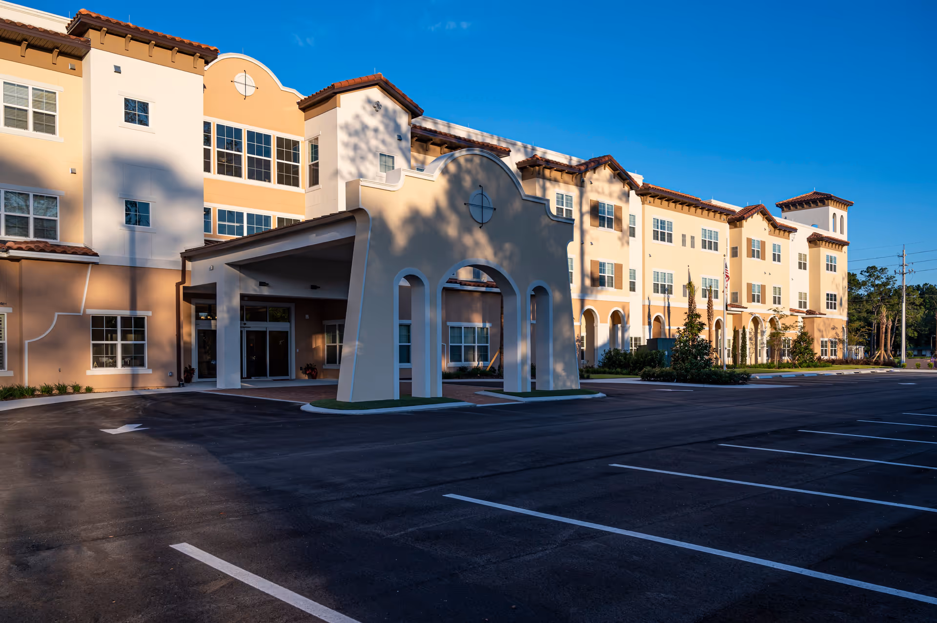 Exterior view of a multi-story senior living facility building with a covered entrance and an empty parking lot under a clear blue sky.