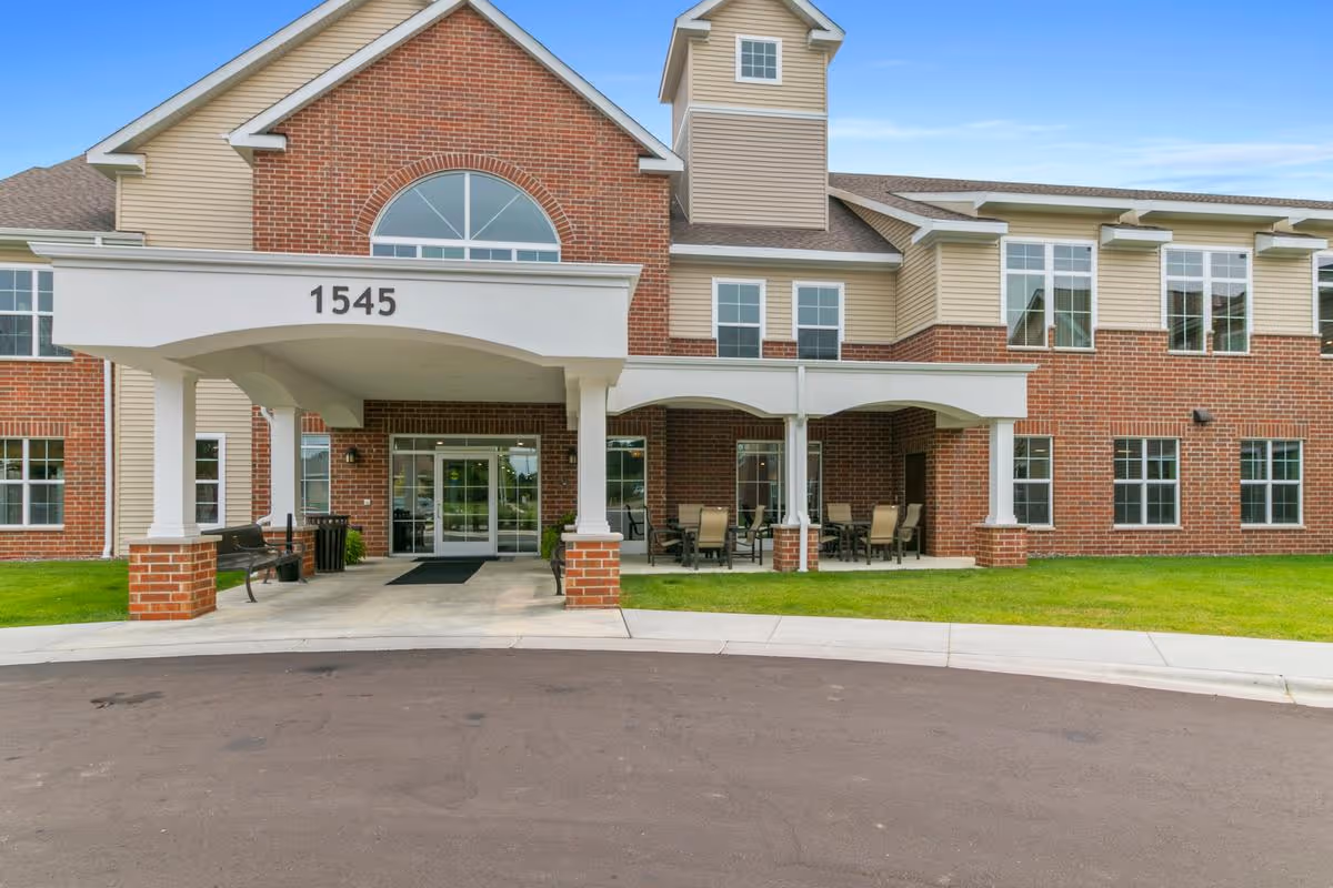Front entrance of a two-story brick senior living building with a covered porte-cochère marked 1545 and outdoor seating by the driveway.