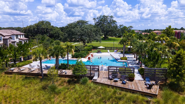 Aerial view of an outdoor swimming pool area surrounded by palm trees and greenery. The pool has people swimming and lounging around it. There are wooden decks with chairs and tables adjacent to the pool. Residential buildings and a large tree are visible in the background under a partly cloudy sky.