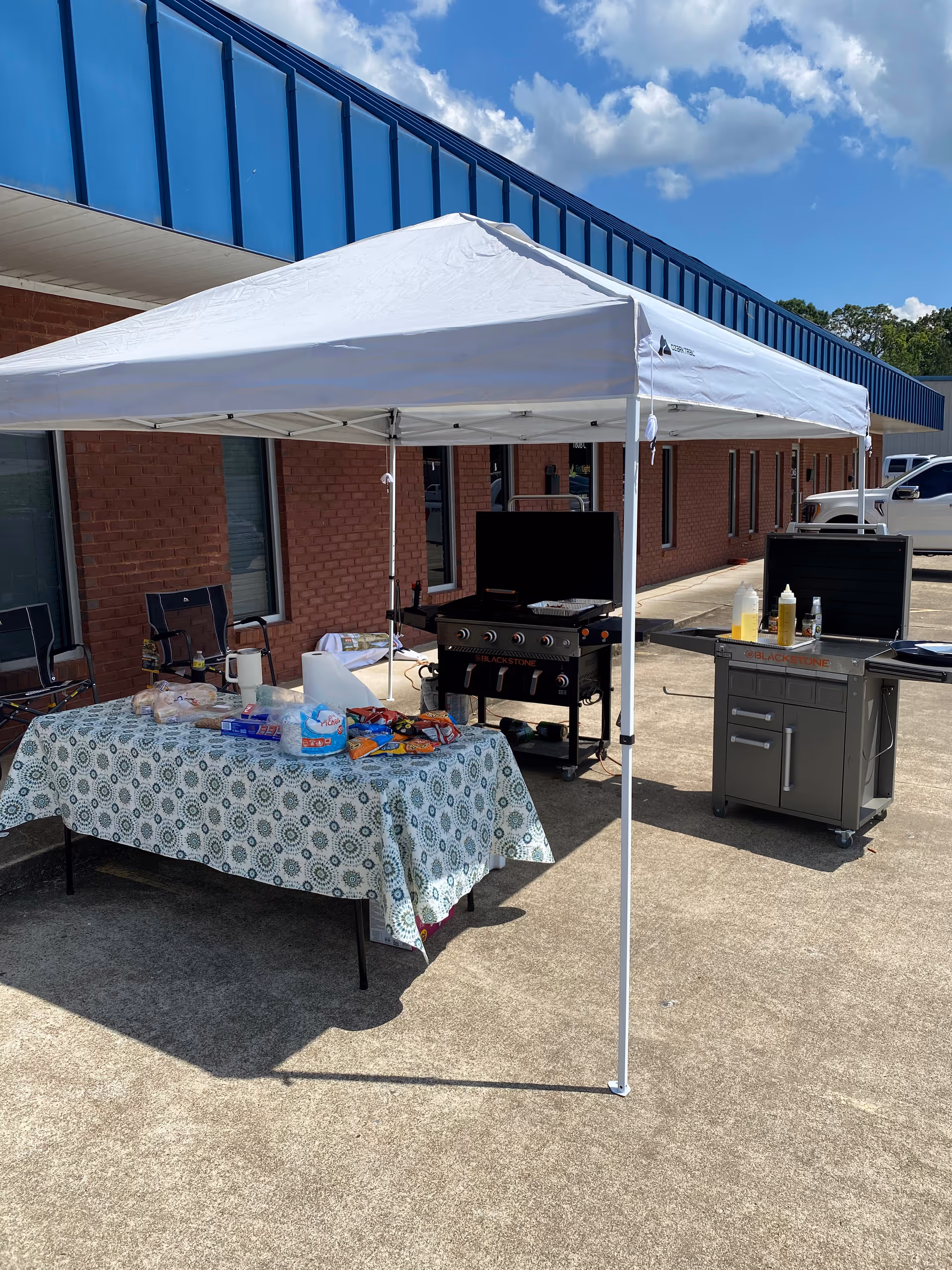 Outdoor setup with canopy, table, grill, and condiments outside a brick building.