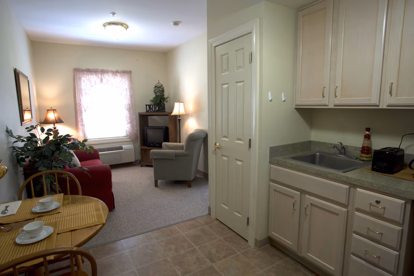 A small living area and kitchenette in a senior living facility. The living area has a red couch, a gray armchair, a wooden TV stand with a small TV, a floor lamp, and a window with lace curtains. The kitchenette features light wood cabinets, a sink, a toaster, and a decorative item on the counter. A small round dining table with two chairs and place settings is visible in the foreground.